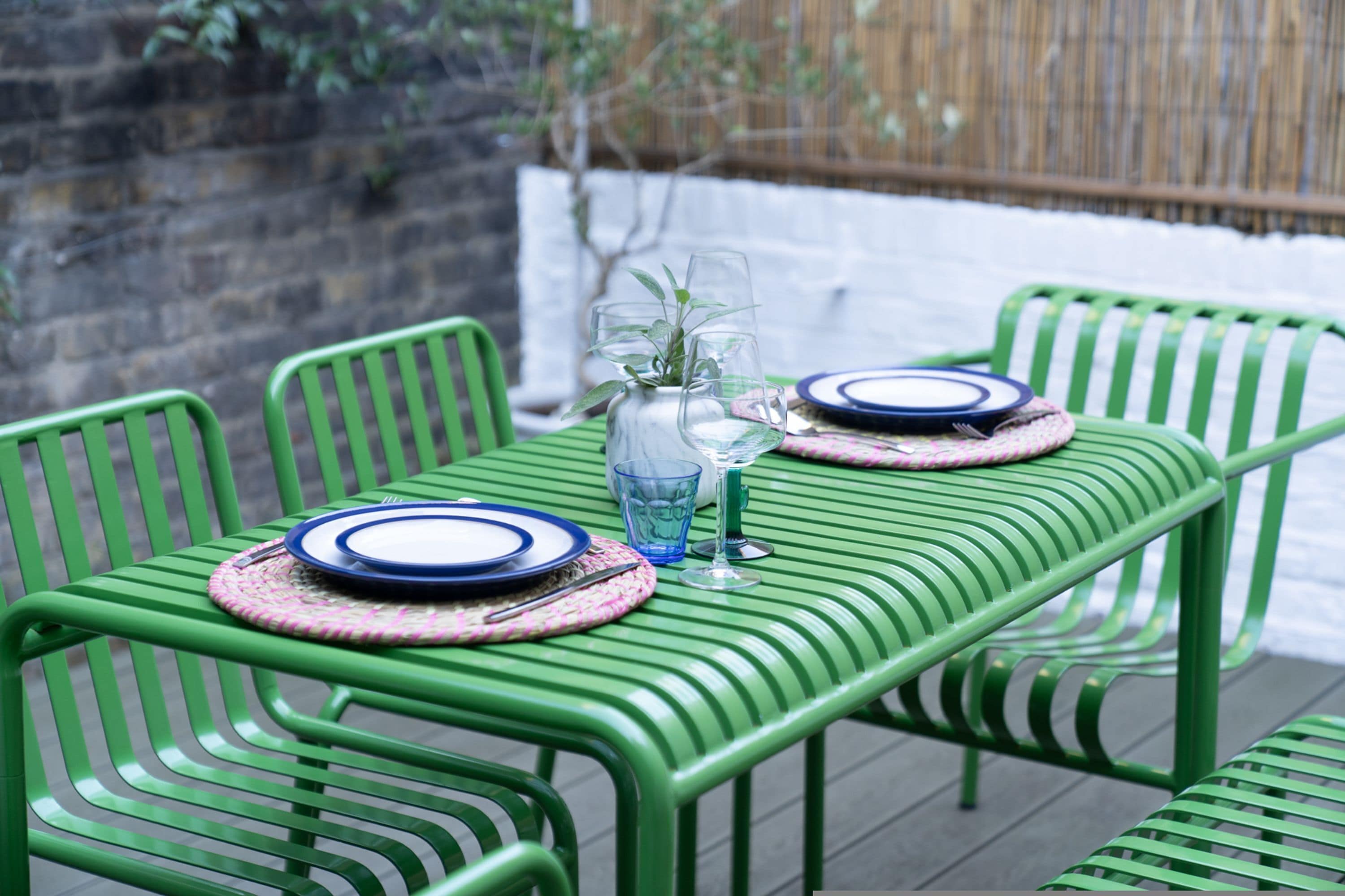 Una mesa de metal verde con platos, cubiertos, un florero con una planta y un vaso.