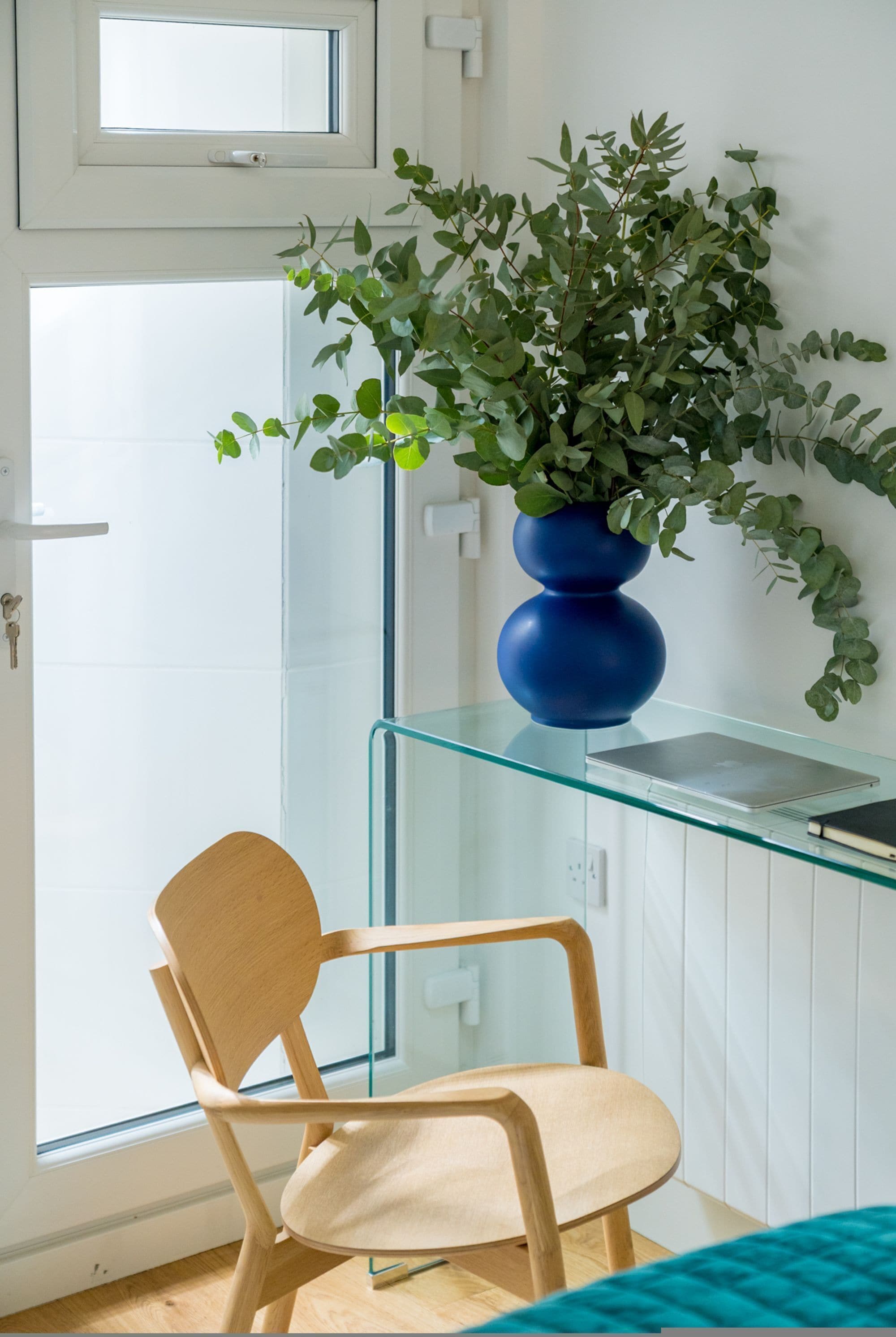 A room with a glass desk, a blue vase with green plants, a wooden chair, and a door.