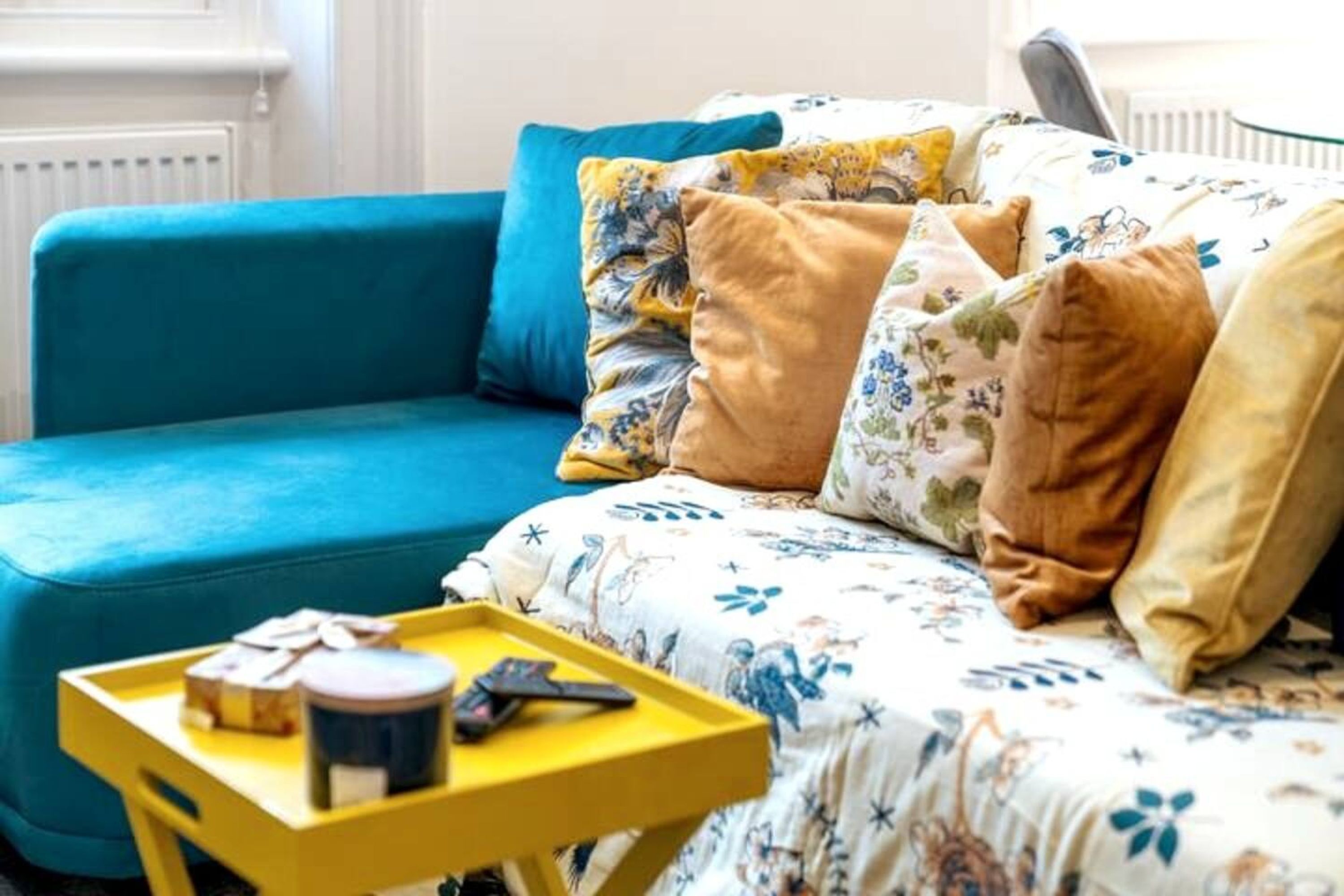Una habitación con un sofá azul, colcha de cama con motivos florales y una mesita auxiliar amarilla con una bandeja.