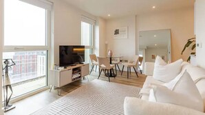 Ein modernes Wohnzimmer mit Flachbildfernseher, einem Esstisch mit Stühlen, einem weißen Sofa und einem großen Fenster.