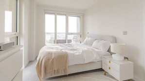 Ein schlicht eingerichtetes Schlafzimmer mit großem Fenster, einem Bett mit weißer Bettwäsche, einem Nachttisch mit Lampe und einer weißen Kommode.