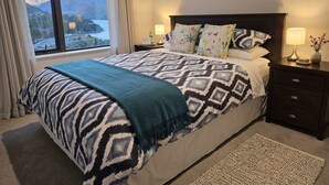 A bedroom with a large bed, two bedside tables, a window with a view of mountains, and two framed pictures on the wall. - Mountain View Exchange (Queenstown)