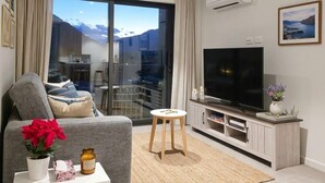 A modern living room with a grey sofa, a small white table with a plant and candle, a TV on a wooden stand, and a large window with a view of mountains. - Mountain View Exchange (Queenstown)