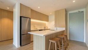 A modern kitchen with a stainless steel refrigerator, wooden cabinets, a white countertop, and a bar stool area. - Mountain View Exchange (Queenstown)