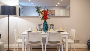 A dining area with a white table, four white chairs, a blue vase with flowers, and a black floor lamp. - Earl s Court Chelsea 2BR Modern Apt Walk to Tube (London)