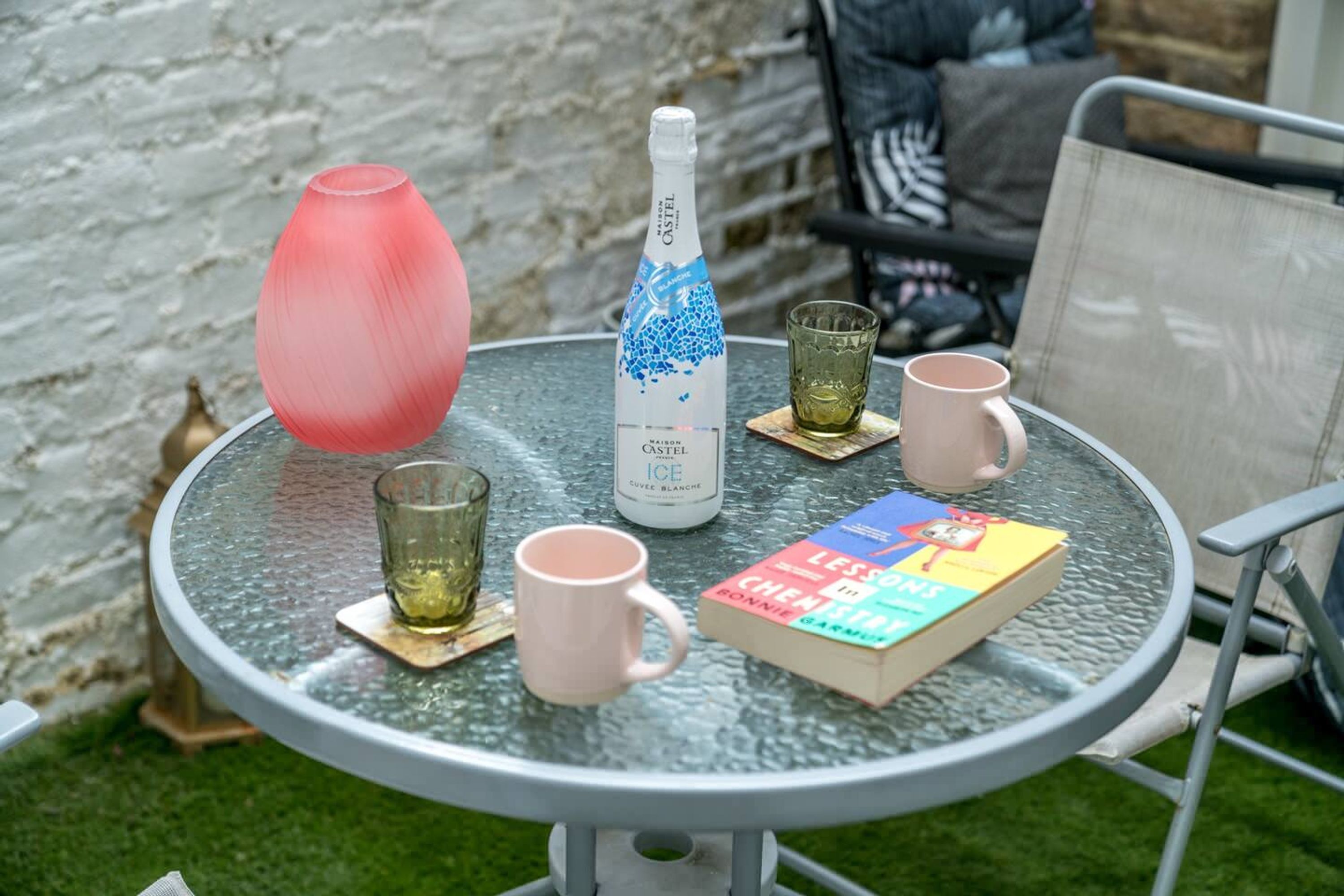 Une table ronde en verre, avec une bouteille de vin, deux verres, un vase rose et un livre.