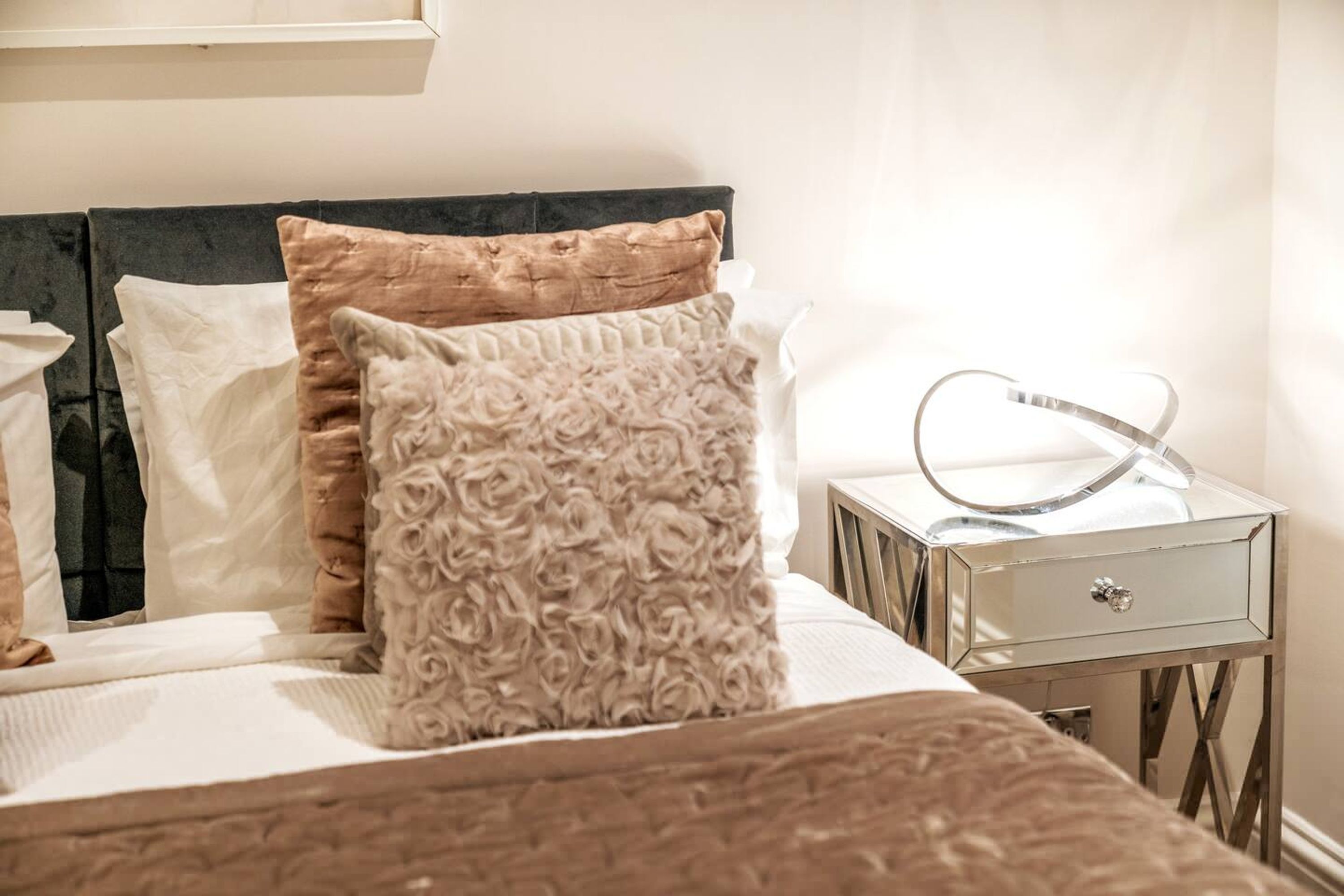 Una cama bien hecha con cabecera, una mesita de noche con espejo y un cojín decorativo.