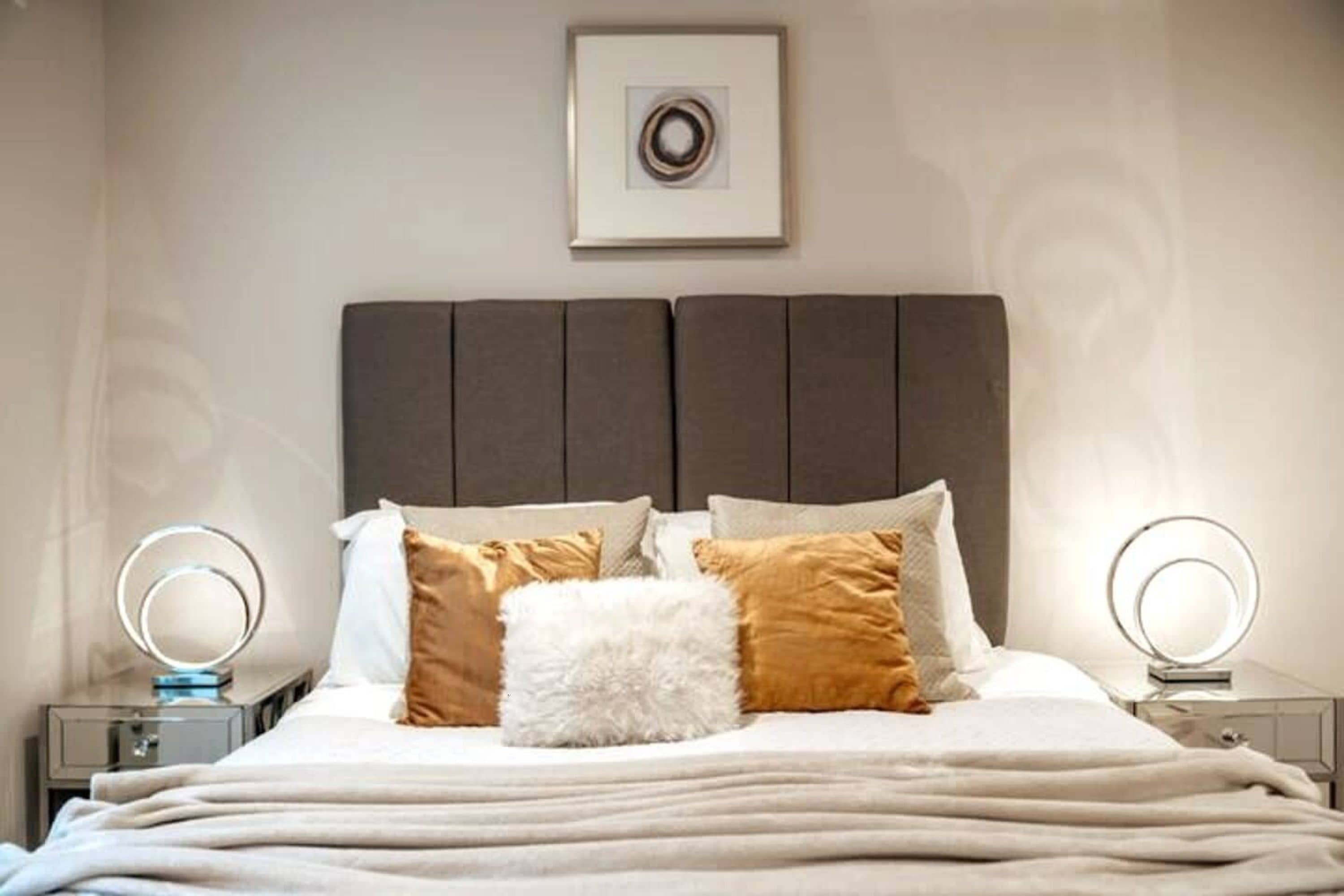 A neatly made bed with a grey headboard, white and brown pillows, and a fluffy white accent pillow. There are two bedside tables with round mirrors.