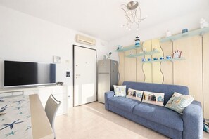 Ein modernes Wohnzimmer mit Fernseher, einem blauen Sofa mit Seekissen, einem Holzschrank mit Regalen und einer Deckenleuchte.