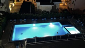 En rektangulär swimmingpool med en omgivande terrass och solstolar, omgiven av byggnader.