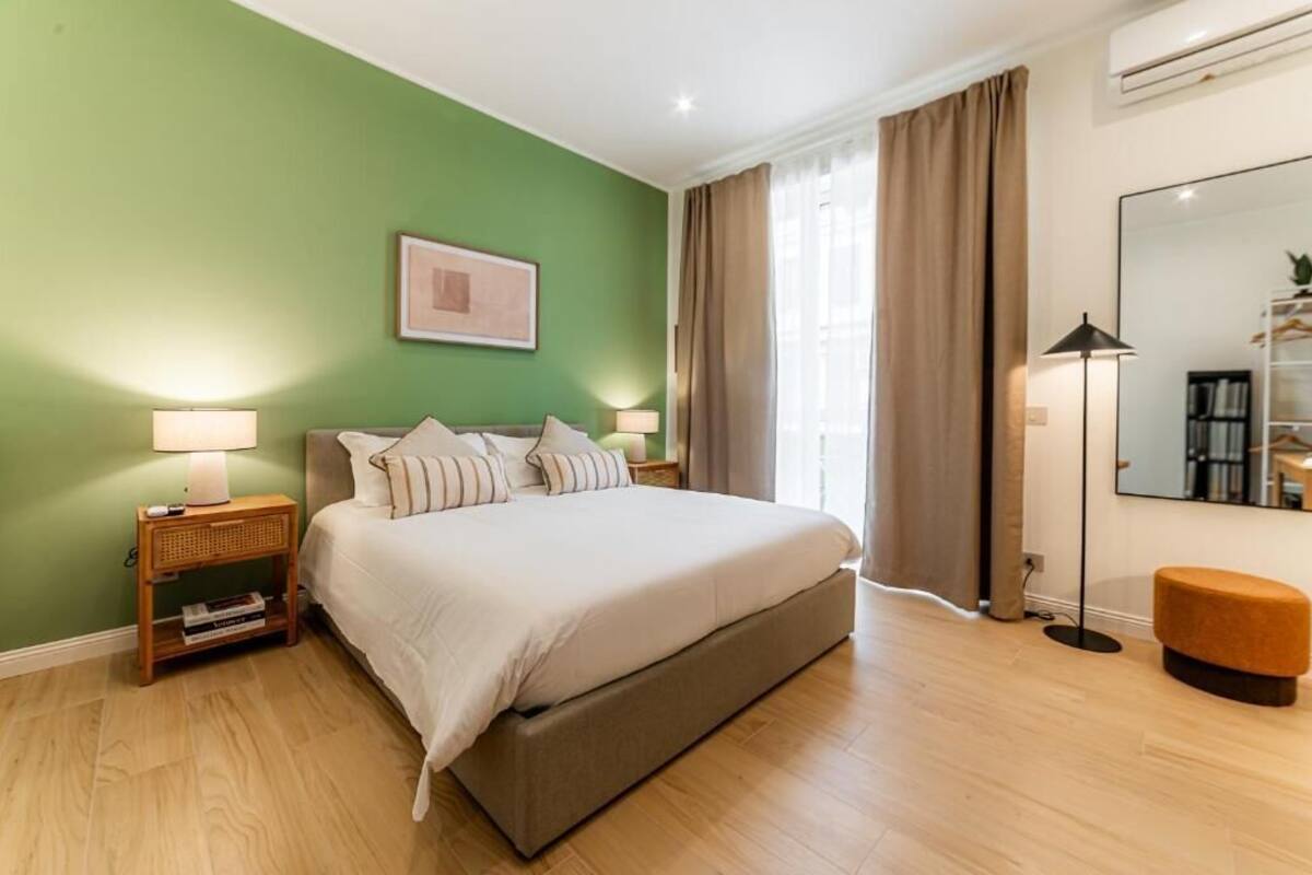 Un dormitorio con cama, lámparas de noche, mesita de noche, espejo y ventana con cortinas.