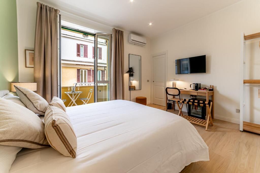 A hotel room with a bed, a desk with a chair, a television, and a balcony with a view of buildings.