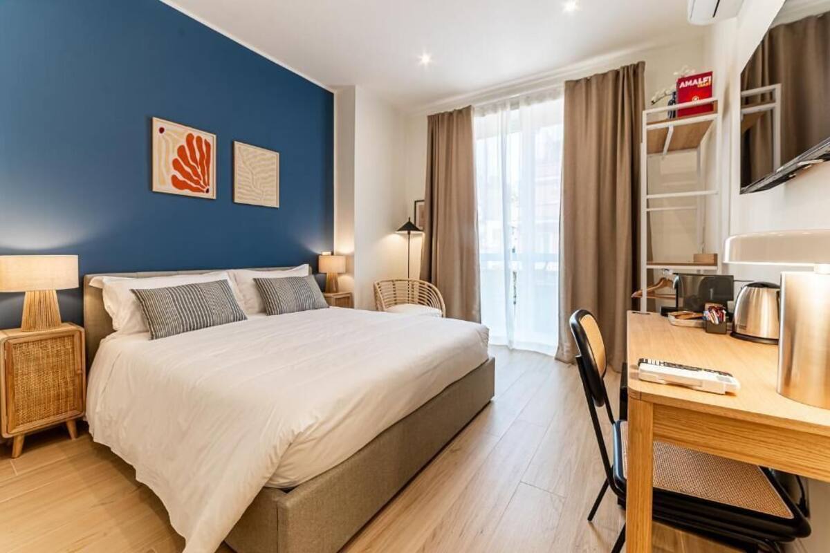 Una habitación de hotel con cama, mesitas de noche, escritorio, silla, lámpara, ventana con cortinas y una obra de arte en la pared.