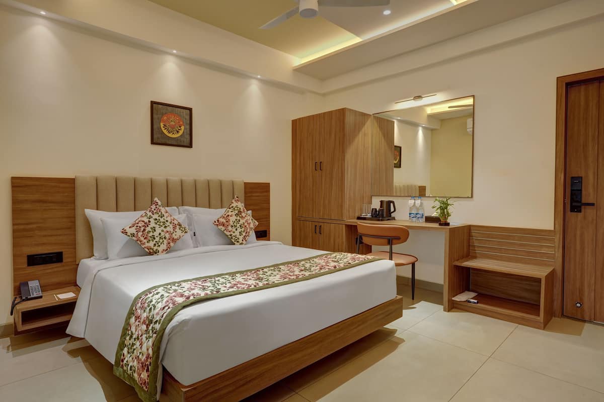 A modern hotel room with a large bed, wooden headboard, bedside tables, a desk with a chair, a mirror, and a wardrobe.