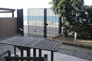 Eine Terrass mit Tisch und Stühlen, ein Tor zum Strand und ein Baum.