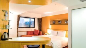 Ein Hotelzimmer mit Bett, Schreibtisch mit Wasserkocher, Stuhl, Fenster mit Vorhängen und Wandregal mit Gegenständen.