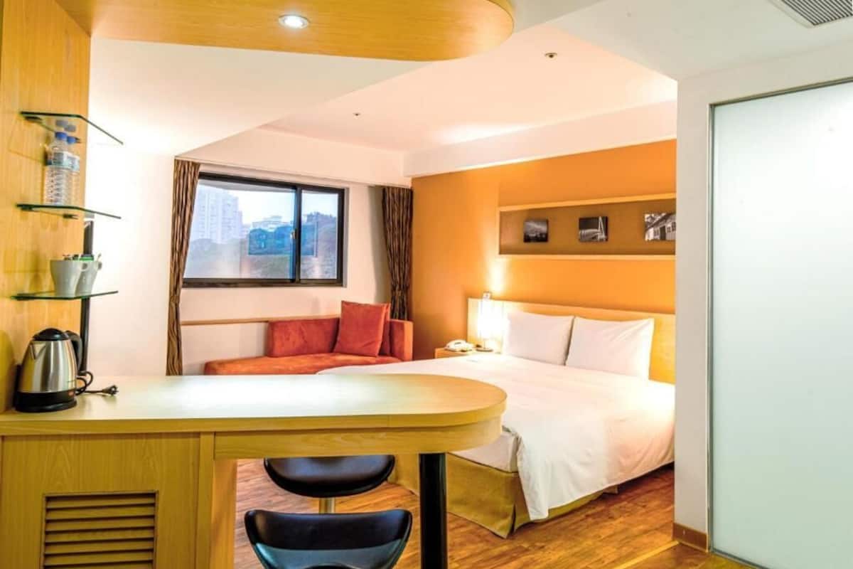 A hotel room with a bed, a desk with a kettle, a chair, a window with curtains, and a wall-mounted shelf with items.