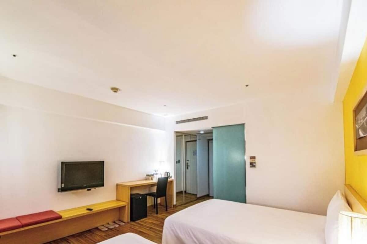 A hotel room with a bed, a desk, a chair, a television, and a red bench.