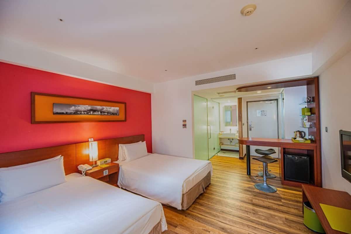 A hotel room with two beds, a desk with a chair, a small kitchenette, and a bathroom visible through an open door.