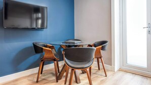 Une pièce avec un téléviseur fixé au mur, une table ronde entourée de chaises et une porte donnant sur un espace lumineux.