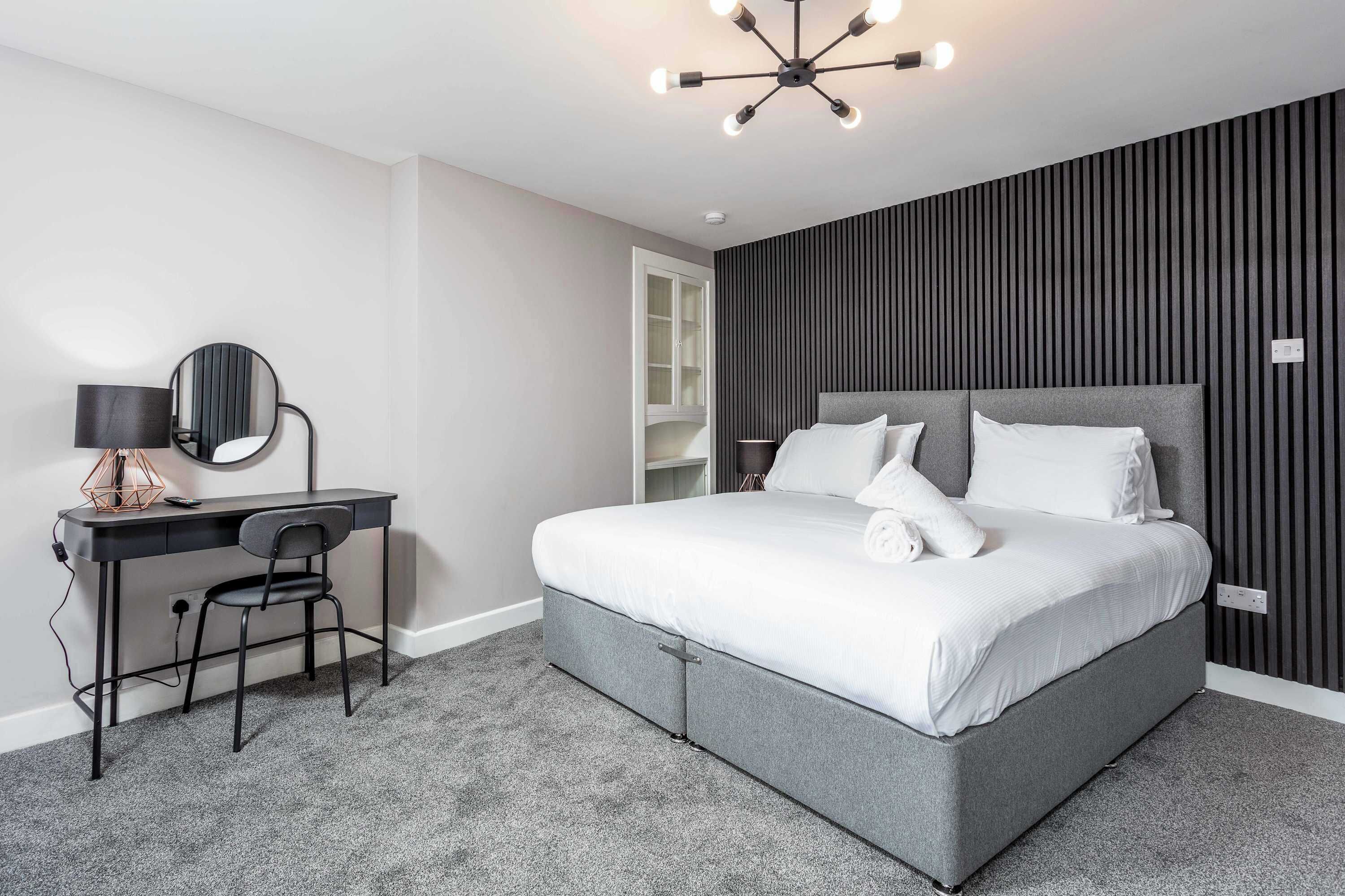 A modern bedroom with a grey bed, a desk with a chair, a round mirror, and a ceiling light fixture.
