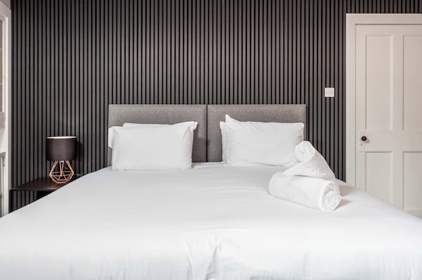 Una cama bien hecha con sábanas blancas, un cabecero gris y una puerta blanca.