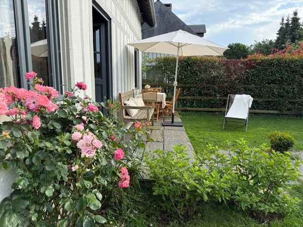 Une terrasse avec des meubles d’extérieur, un parasol blanc et un jardin où poussent des roses roses.