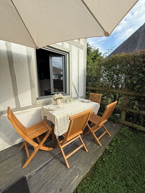 Eine Terrasse im Freien mit einem Tisch für zwei Personen, Holzstühlen, einer weißen Tischdecke und einem Fenster mit Blick auf einen Garten.