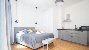 Een slaapkamer met een bed, nachtkastje, bankje en een kitchenette met een magnetron.