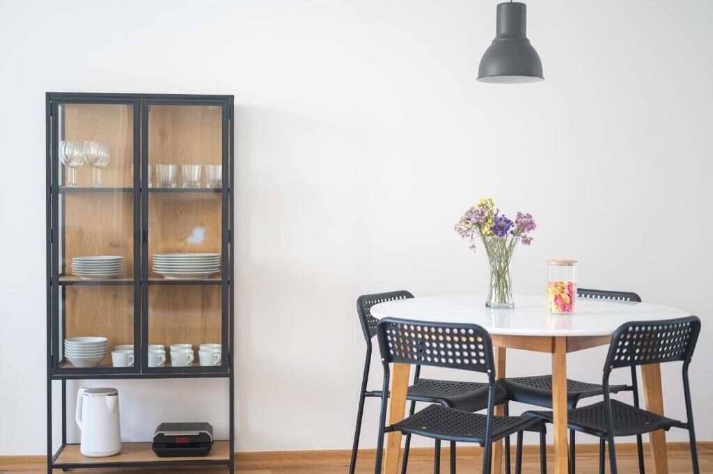 Un comedor con una mesa redonda, cuatro sillas, un florero con flores, un armario con vajilla y un hervidor.