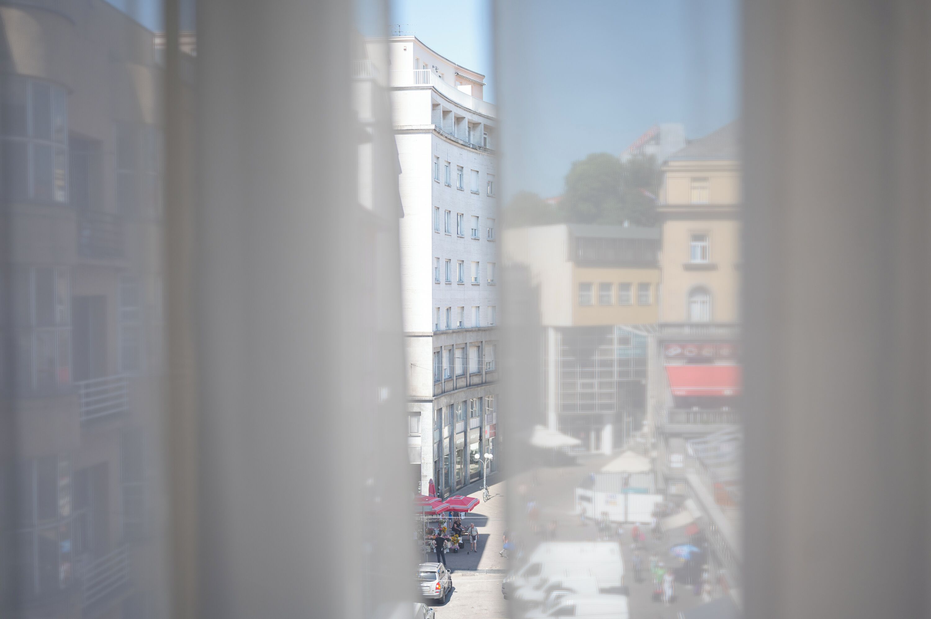 A view through a window of a multi-story building with balconies, a street with parked cars, and a clear sky.