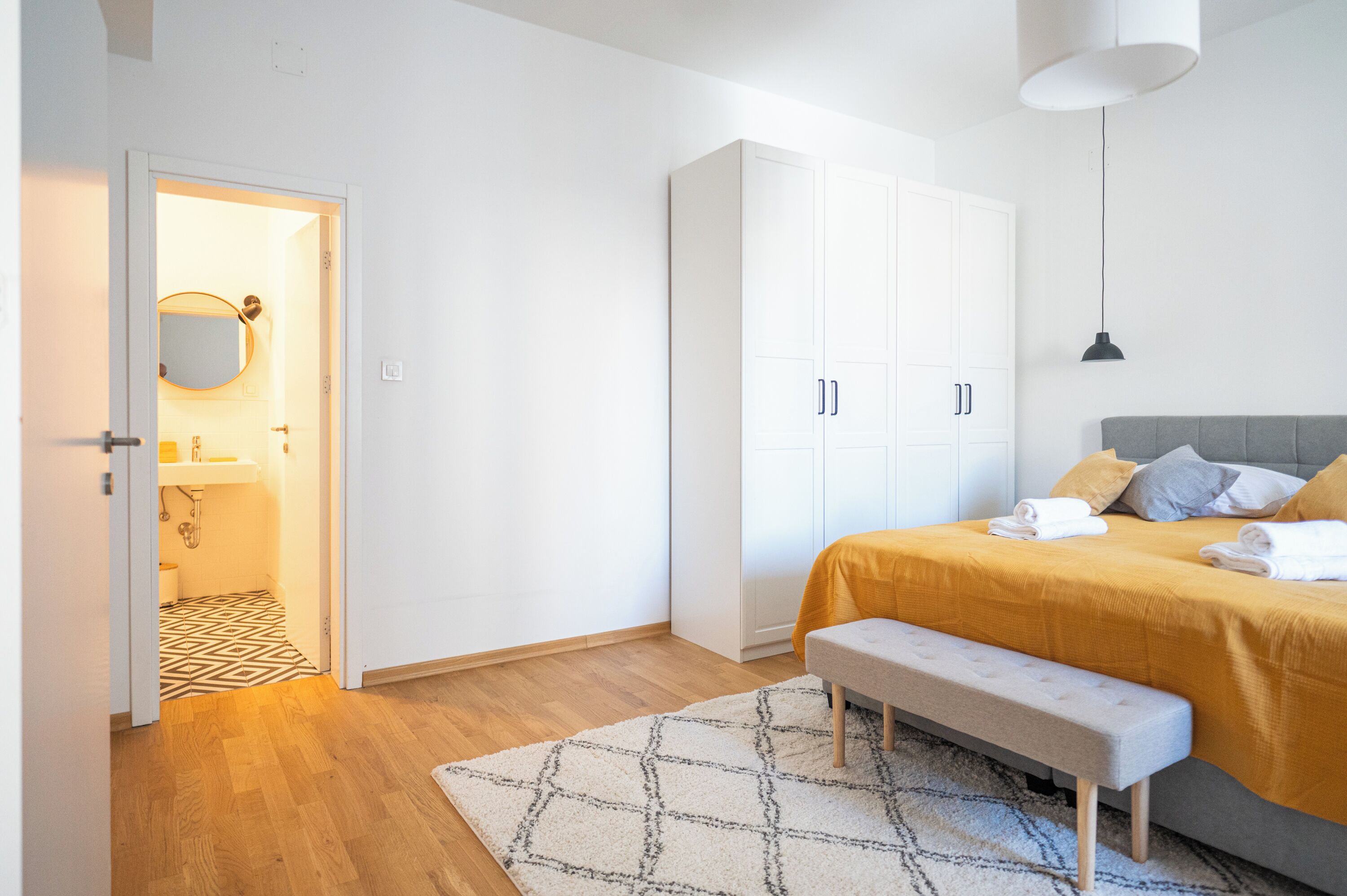 A bedroom with a bed, bench, wardrobe, and a bathroom visible through an open door.