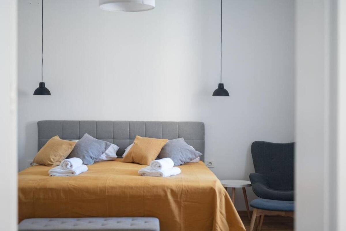 Una cama bien hecha con cabecera gris, dos lámparas colgantes negras y una pequeña mesa redonda con una silla a un lado.