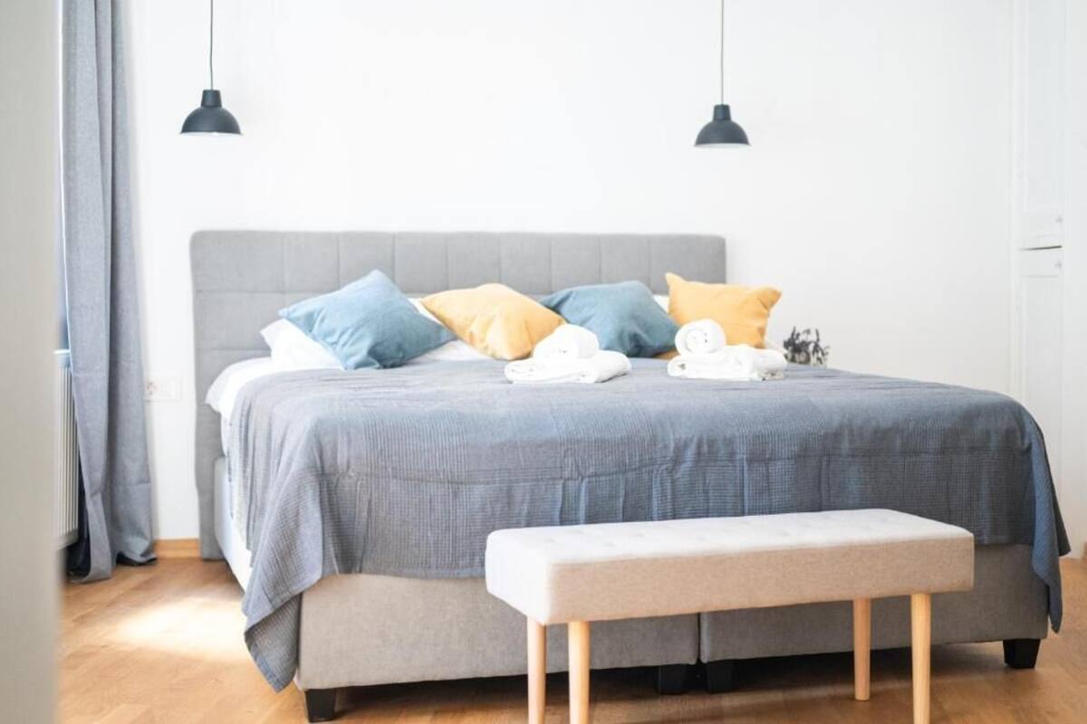 Un dormitorio con cama, almohadas, un banco y dos lámparas colgantes.
