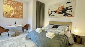 Ein modernes Hotelzimmer mit einem Bett, einem runden Esstisch, zwei Stühlen, einem großen abstrakten Gemälde und einem Nachttisch mit einer Lampe.
