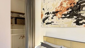 Ein Schlafzimmer mit Rattan-Kopfstütze, einem Bett mit grau und weißen Kissen, einem an der Wand hängenden abstrakten Gemälde und einem Kleiderschrank mit darin befindlicher Kleidung.