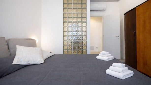 A bedroom with a grey sofa, a wooden wardrobe, and a glass partition wall. - Sonnino 17 Luxury Apartment (Cagliari)