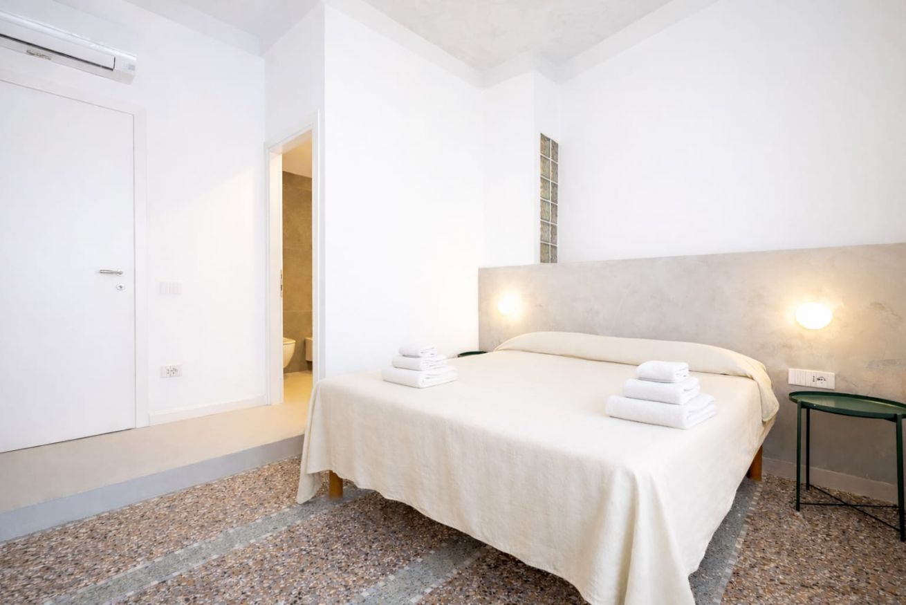 Una camera d'albergo con un letto, un comodino, una lampada a parete e una porta.