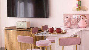 Une cuisine moderne aux accents rose et or, dotée d’un téléviseur à écran plat et d’une machine à café.