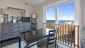 Quarto com mesa de jantar, cadeiras, televisão em um móvel, estante e vista para o mar por grandes janelas.