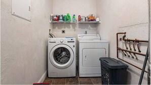 Uma lavanderia com máquina de lavar, secadora, prateleiras com produtos de limpeza e suporte para mangueira.