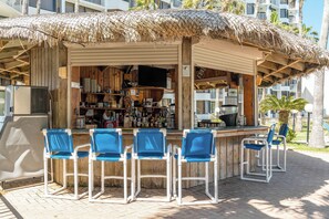 A beachside bar with a thatched roof, wooden walls, and a counter with various bottles and a television. There are four blue chairs and a table in front of the bar. - Saida Royale 145 - Premier Location with Breathtaking Ocean Views. The Only Thing Missing is You! (South Padre Island)
