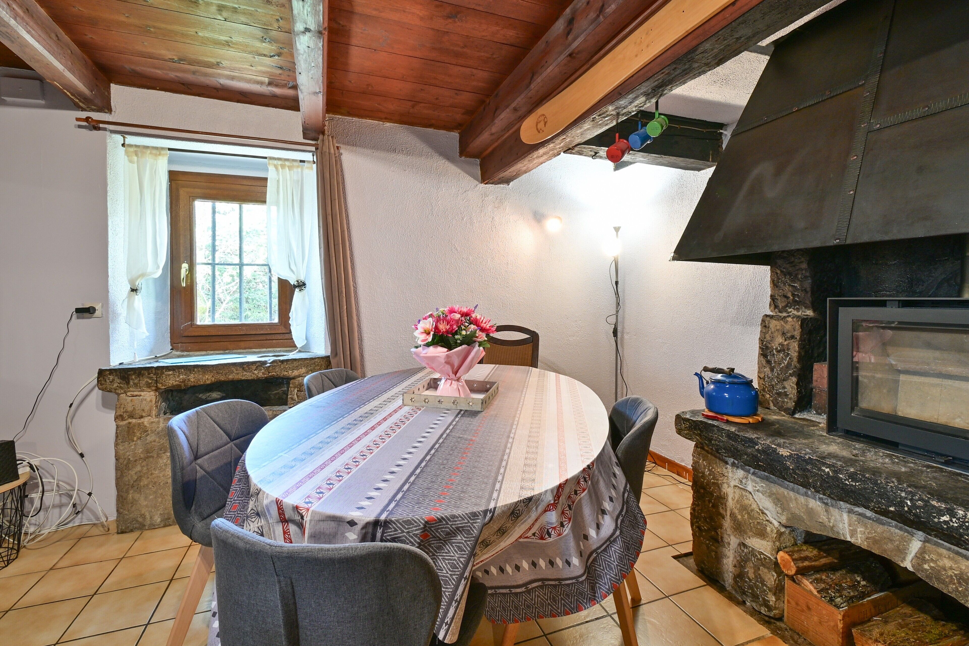 Un comedor con una mesa redonda, sillas, un centro de mesa con flores, una chimenea de piedra y un ventanal con cortinas.