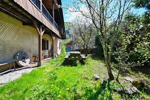 A rustic wooden building with a balcony, a wooden bench, and a picnic table in a lush garden.