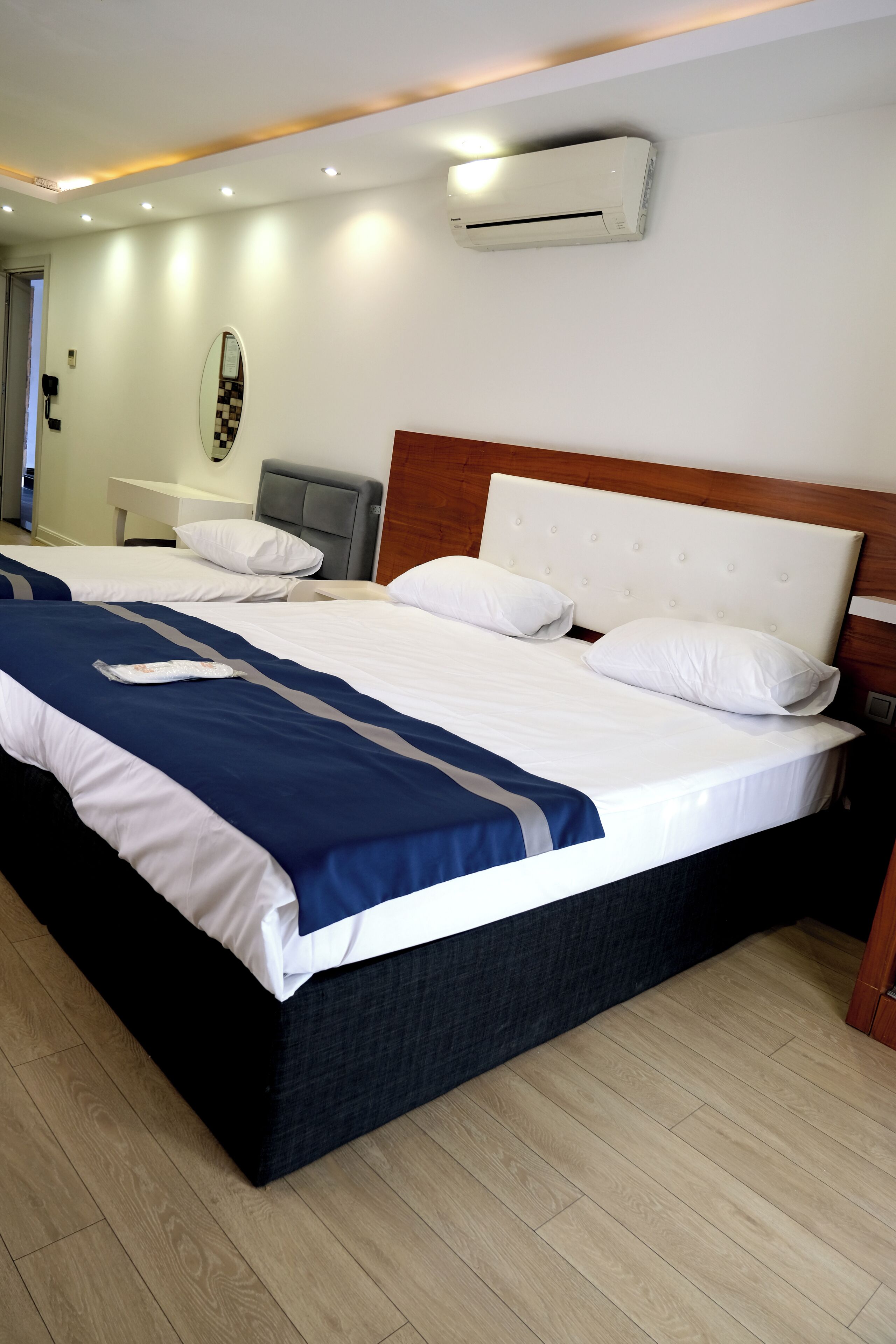 Una cama doble con colcha blanca y azul, cabecera de madera y un equipo de aire acondicionado en la pared.