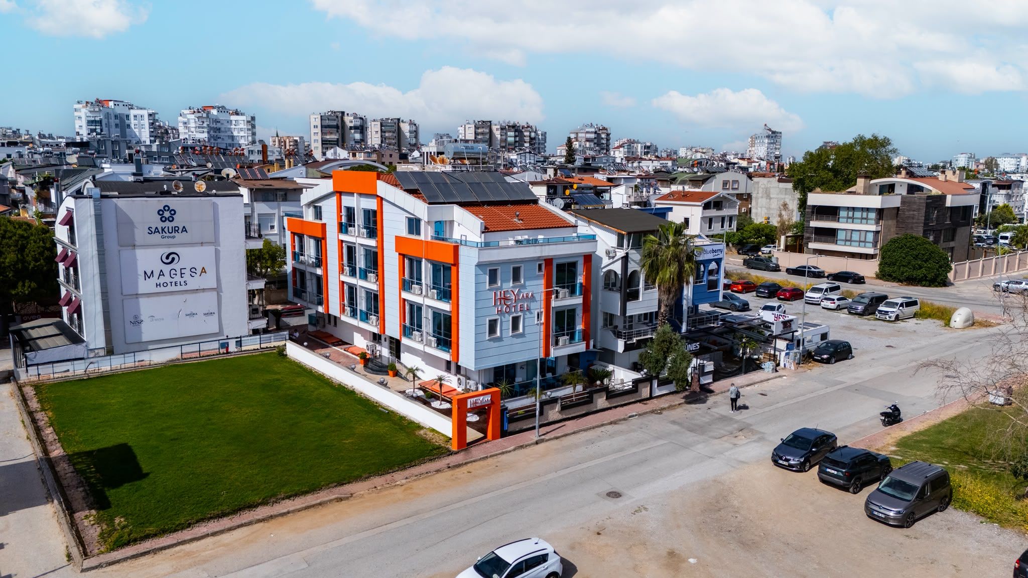 Un hotel de varios pisos con un diseño moderno, que destaca por su fachada azul y naranja, un jardín en la azotea y un estacionamiento con varios coches.