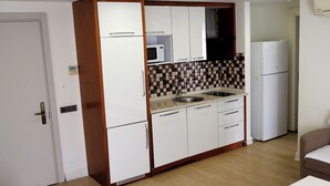A compact kitchen with a microwave, sink, and refrigerator, a bed with a headboard, and a small table.