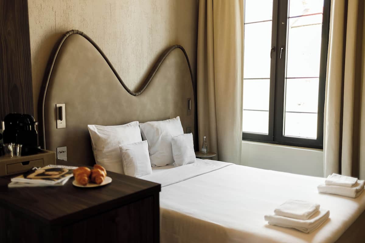 A hotel room with a bed, pillows, a bedside table with a tray of croissants, a coffee machine, and a window with curtains.