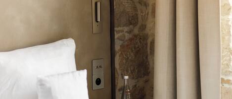 A bed with white linens, a stone wall, a small bedside table with a bottle, and a curtain.