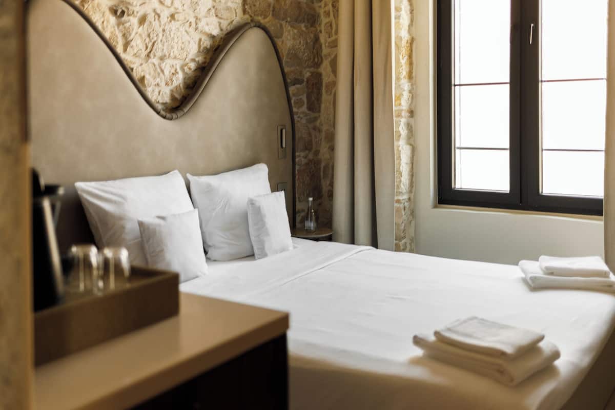 A hotel room with a stone wall, a bed with white linens, a wooden nightstand, and a window with curtains.